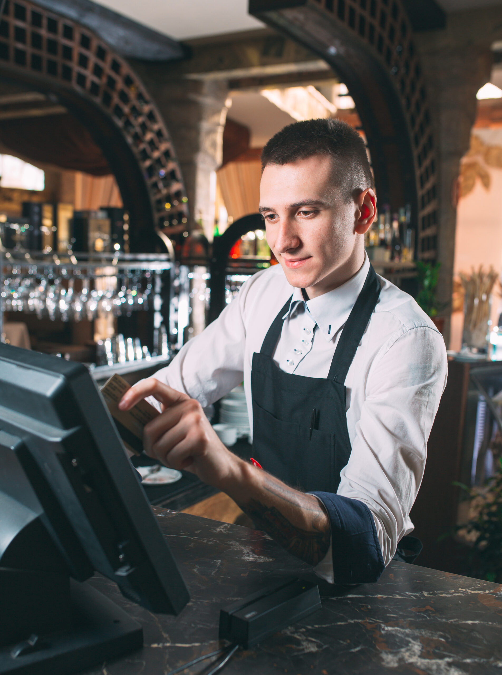 small business, people and service concept - happy man or waiter in apron at counter with cashbox working at bar or coffee shop